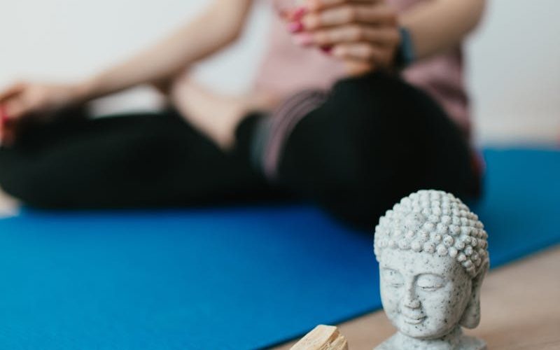 Selective focus of crop faceless young female in casual wear meditating in lotus pose with white sage incense near Buddha head sculpture amethyst crystal and palo santo wood in stone bowl